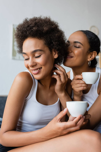 happy lesbian african american couple holding cups of coffee while smiling at home - Foto, immagini