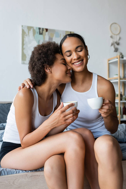 cheerful lesbian african american couple holding cups of coffee while smiling at home - Photo, image