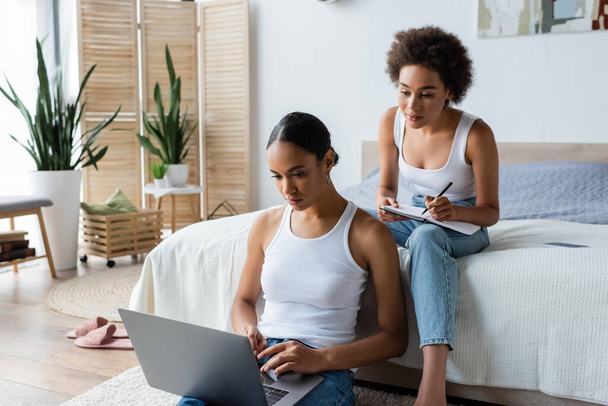 young african american woman with notebook looking at laptop near girlfriend in bedroom - Φωτογραφία, εικόνα