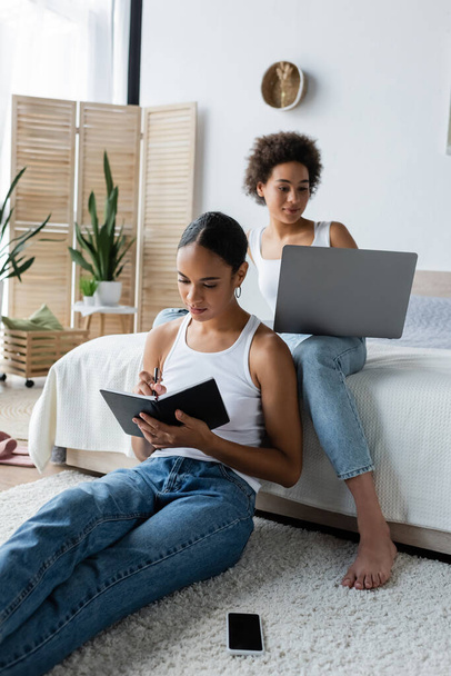 afro-amerikai nő írásban notebook közelében barátnője dolgozik otthonról laptop - Fotó, kép