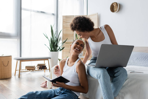 happy african american woman with laptop looking at cheerful girlfriend holding notebook - Photo, Image