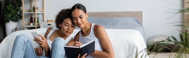 lesbian african american woman holding smartphone and looking at notebook in hands of girlfriend, banner  - Foto, Imagen