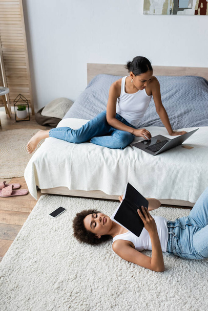 encaracolado afro-americano mulher deitada no tapete e olhando para notebook perto namorada usando laptop na cama - Foto, Imagem