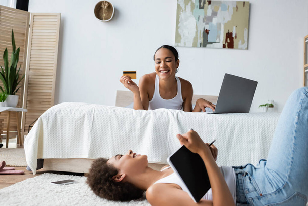 jovem afro-americana deitado no tapete e olhando para a namorada feliz segurando cartão de crédito ao usar laptop  - Foto, Imagem