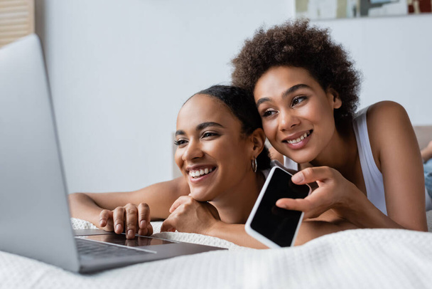 feliz africano americano lesbiana mujer celebración smartphone con pantalla en blanco mientras busca portátil cerca de novia  - Foto, Imagen