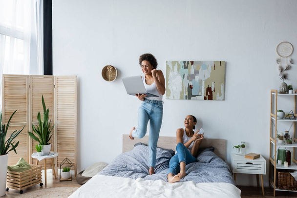 excited african american lesbian woman holding laptop while standing on bed near smiling girlfriend   - Photo, Image