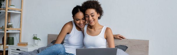 african american lesbian couple looking at laptop while watching movie in bedroom, banner - Foto, afbeelding