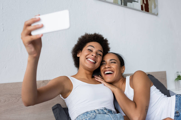 幸せなアフリカ系アメリカ人レズビアンカップル取ります自画撮りスマートフォンでベッドルーム - 写真・画像