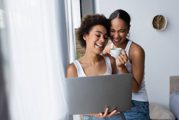 cheerful african american lesbian woman using laptop near happy girlfriend holding cup of coffee  - Foto, Imagem