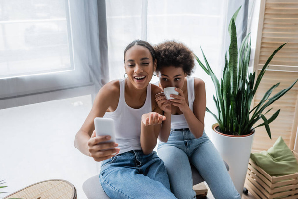 happy african american lesbian woman talking during video chat on smartphone while girlfriend drinking coffee - Фото, изображение