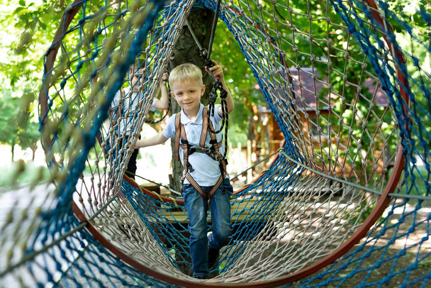 Portrait of a little boy in climbing gear in a rope park, holding a rope with a carbine - 写真・画像
