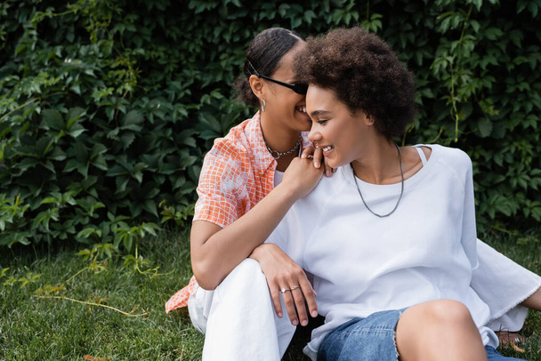 cheerful african american lesbian woman in sunglasses hugging happy girlfriend while sitting on lawn  - Zdjęcie, obraz
