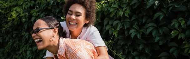 happy african american lesbian woman embracing excited girlfriend in sunglasses laughing in green park, banner - Фото, изображение