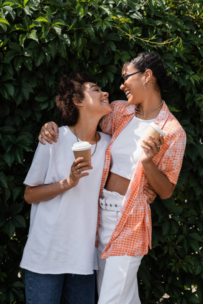 joyful african american lesbian woman in sunglasses embracing cheerful girlfriend and holding coffee to go - Foto, imagen