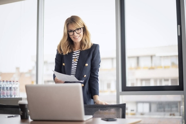 Shot of a confident mature businesswoman having video call while standing in a modern office and holding document in her hand. Professional female wearing eyeglasses and blazer. - Fotoğraf, Görsel