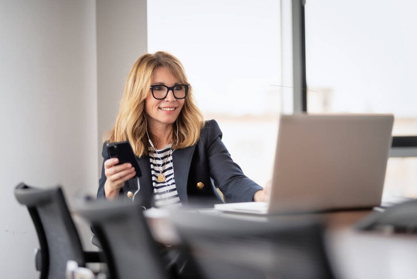 Femme d'affaires d'âge moyen souriante utilisant un smartphone et des écouteurs assis au bureau et ayant un appel vidéo.  - Photo, image