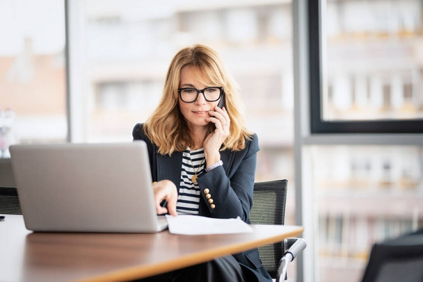 Middle aged businesswoman using laptop and making a call while sitting at desk at the office. Executive businesswoman wearing eyeglasses and blazer. - 写真・画像