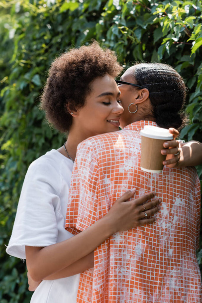 happy african american lesbian woman hugging girlfriend and holding coffee to go - Zdjęcie, obraz