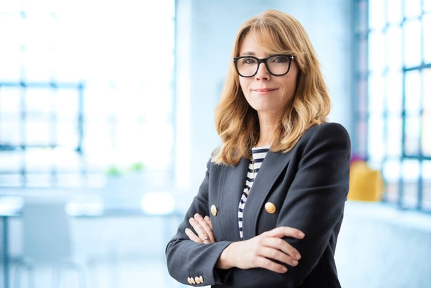 Mature female with arms crossed. Confident businesswoman looking at camera and smiling while standing at the office. Attractive woman wearing eyewear and blazer.  - Foto, Imagem