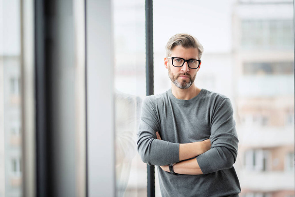 Thinking businessman wearing sweater and eyewear while standing with arms crossed at glass wall in the office. - Foto, afbeelding