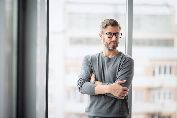 Ajatteleva liikemies yllään pusero ja silmälasit seisoessaan kädet ristissä lasiseinällä toimistossa. - Valokuva, kuva