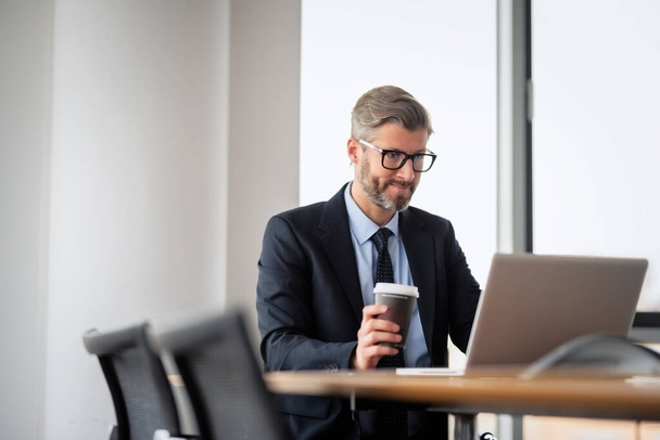 Середній дорослий бізнес-професіонал сидить за столом з ноутбуком і п'є каву під час роботи в сучасному офісі
. - Фото, зображення