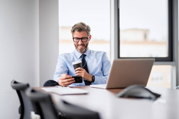 Mid adult business professional sitting at table with laptop and talking on mobile phone. Businessman having video call while working in office. - Photo, Image
