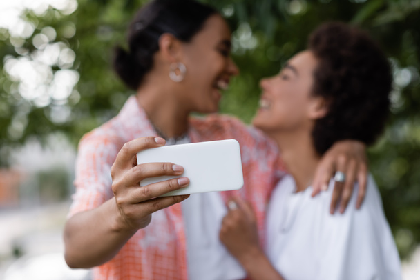positive african american lesbian couple taking selfie on blurred background  - Zdjęcie, obraz
