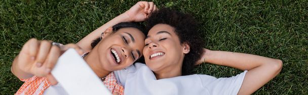 top view of joyful african american lesbian woman holding smartphone and taking selfie with girlfriend lying on grass, banner - Фото, изображение