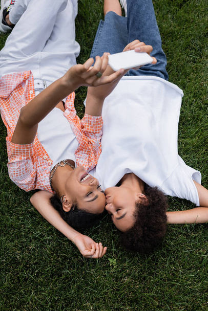 top uitzicht van gelukkig Afrikaans amerikaans lesbisch vrouw holding smartphone en het nemen van selfie met vriendin liggend op gras  - Foto, afbeelding
