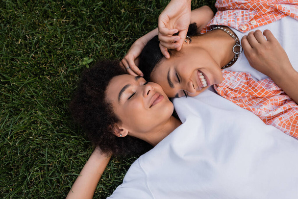 top view of happy african american lesbian women lying on grass  - Zdjęcie, obraz