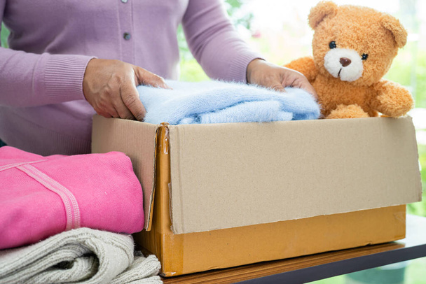 Κρατώντας κουτί δωρεά ενδυμάτων με μεταχειρισμένα ρούχα και κούκλα στο σπίτι για την υποστήριξη της βοήθειας για τους φτωχούς ανθρώπους στον κόσμο. - Φωτογραφία, εικόνα