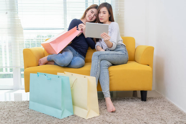 Two asian young woman sitting on the yellow sofa using digital tablet  shopping online near window and shopping bag - Фото, изображение