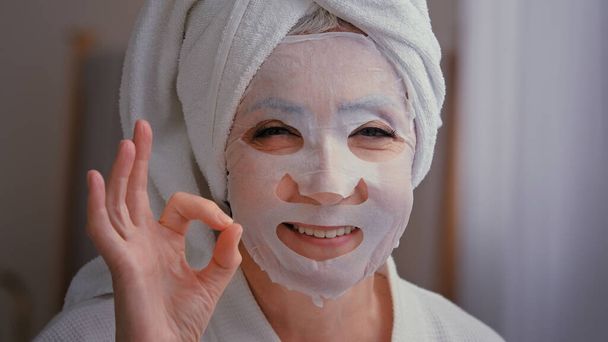 Close up stary starszy Dojrzałe biały kobieta z skóra pielęgnacja tekstylny skóra maska na kobiecej twarzy pokazując ok dobre piękno kosmetologii procedury 60s lady wrap ręcznik na głowie umieścić nawilżający lifting kosmetyk - Zdjęcie, obraz