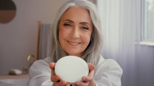 50 'li yaşlarda, mutlu, beyaz saçlı, olgun bir kadın banyoda yaşlı, dişlek, 60' lı yaşlarda, kozmetik karşıtı, organik, mineral kremli cilt bakımını nemlendiren bir kavanoz doğal güneş kremi gösteriyor. - Fotoğraf, Görsel