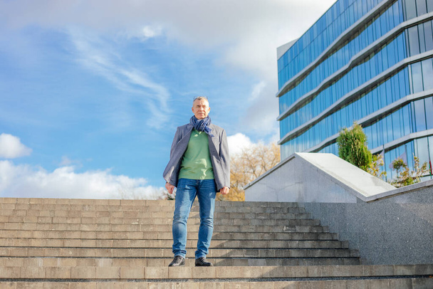 Portrait of middle-aged man businessman with short hair wearing grey jacket, blue scarf, jeans, green T-shirt, going down concrete stairs near modern buildings, shrubs in city outdoors in autumn. - Fotó, kép