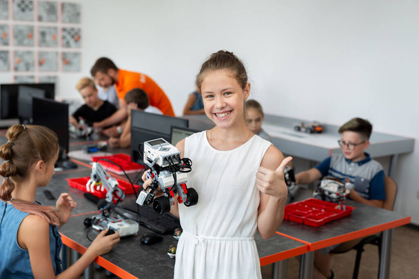 Portrait of a happy schoolgirl girl in a robotics class, she holds a robot assembled from plastic parts programmed on a computer - Fotoğraf, Görsel