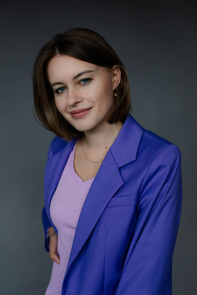 Portrait of young smiling amazing blue-eyed woman businesswoman with short dark hair wearing blue jacket, holding hand on waist, looking at camera posing on grey background. Studio, vertical. Fashion. - Foto, immagini