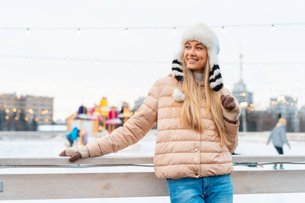 Beautiful lovely middle-aged girl blonde hair warm winter jackets knitted glove stands ice rink background Town Square. Christmas mood lifestyle Happy holiday woman walk snowy day Winter leisure - Foto, immagini