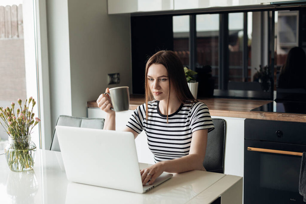 Сконцентрированная, серьезная темноволосая деловая женщина, сидящая на кухне, работающая на белом ноутбуке и пьющая кофе возле большого окна. Беспроводные технологии и интернет в независимом домашнем офисе - Фото, изображение