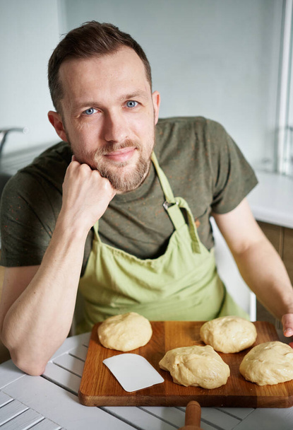 Portret van een blanke bakker in een groen schort uniform lachend naar de camera kijkend. Portret van knappe bakker thuis zittend aan tafel met deeg. Hoge kwaliteit verticale foto - Foto, afbeelding