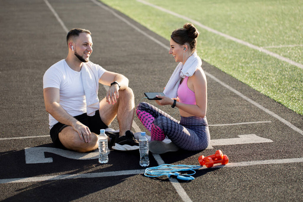 Sporty young boy and woman sit on the treadmill at the stadium in the summer after a run with a phone, headphones and sports bottles - Foto, Imagem