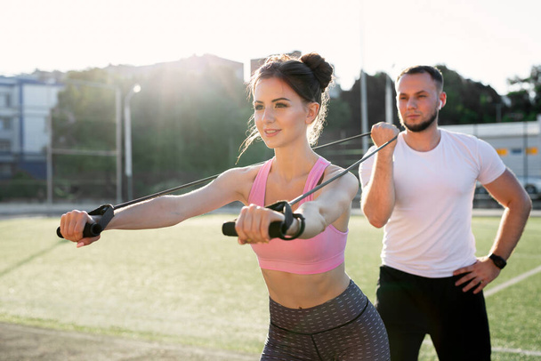 Close-up of young sports people training at the stadium in the summer, a woman doing exercises with an expander and a man helping her - Photo, Image
