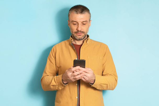 Mature caucasian man using cellphone, texting via smartphone, communicating online standing over blue background. Communication and technology concept - Photo, Image