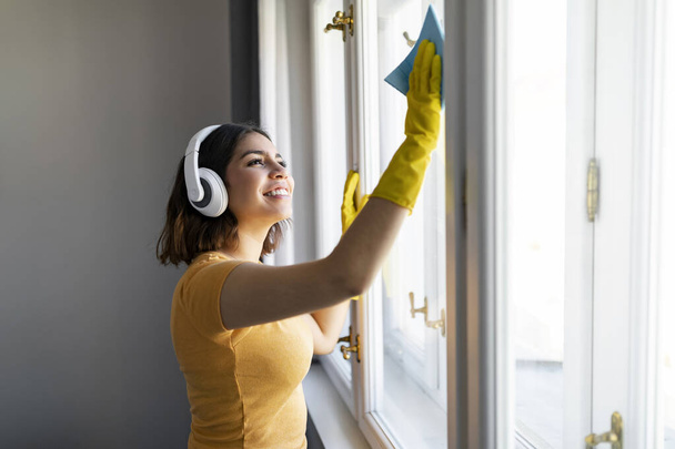 Χαμογελώντας νεαρή αραβίδα γυναίκα πλυντήριο παράθυρο στο σπίτι με Rag, χαρούμενη γυναίκα της Μέσης Ανατολής φορώντας ασύρματα ακουστικά καθαρισμού σπίτι, ακούγοντας μουσική, ενώ κάνοντας εγχώριες χορωδίες, Αντιγραφή χώρου - Φωτογραφία, εικόνα