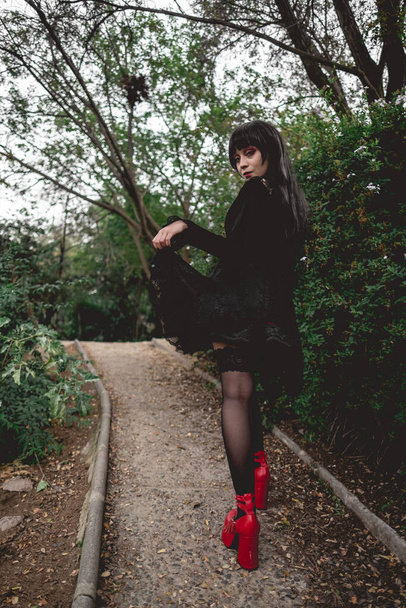 Young and skinny hispanic goth girl with black dress, pantyhose and red platforms shoes walking in a path in the forest - Photo, Image