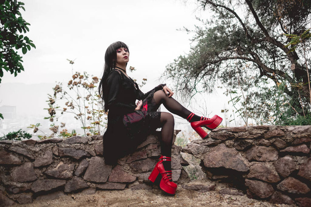Menina gótica hispânica jovem e magro com vestido preto, meia-calça e sapatos de plataformas vermelhas sentados em uma pequena parede de pedra na floresta - Foto, Imagem