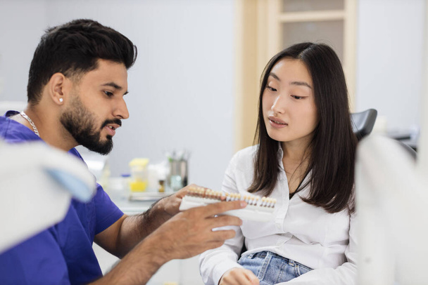 Attractive male dentist checking and selecting color of young asian woman teeth. Visual method of subjective perception color of filling by a doctor. Dentist choosing color of filling from palette - Foto, Imagem