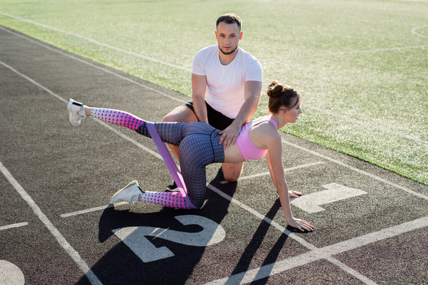 Coach plays sports with a young girl at the stadium. Leg exercise with elastic band - Foto, immagini