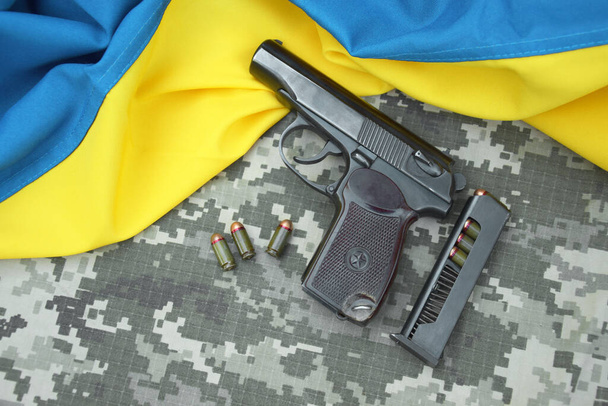 Ένα πιστόλι Makarov με φυσίγγια στο φόντο ενός στρατιωτικού pixel καμουφλάζ και τη σημαία της Ουκρανίας. Ο πρωθυπουργός είναι πιο κοντός. Πόλεμος στην Ουκρανία. - Φωτογραφία, εικόνα